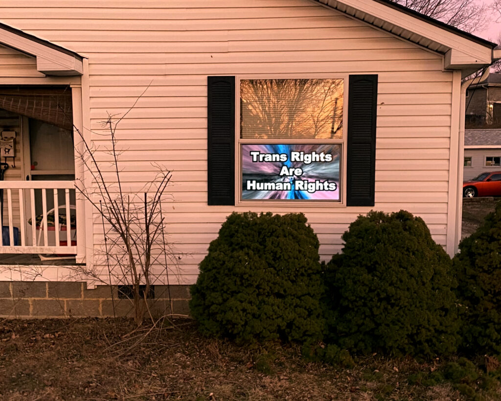 The Lighthouse - a home with a 50 inch screen mounted in the front-facing window. The screen is displaying a blue, pink, and white vortex motion graphic that says "Trans Rights Are Human Rights."